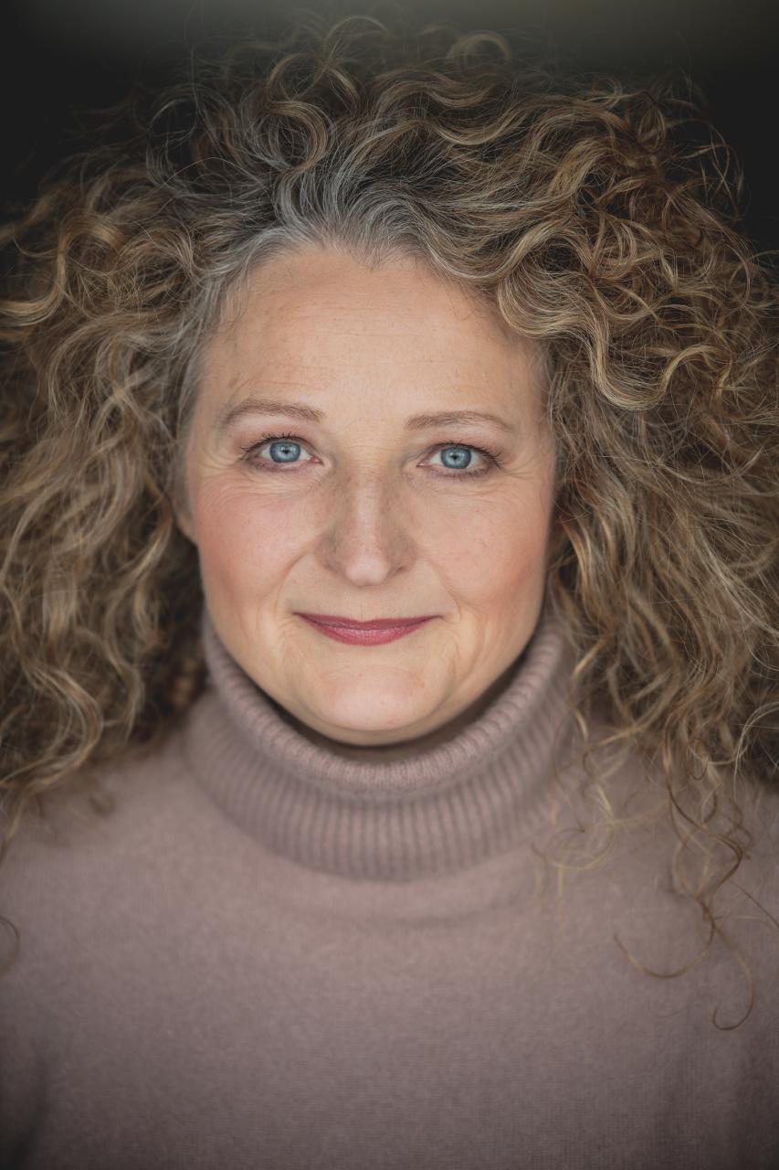 Mona Deibeles Kopf und Oberkörper sind zentral im Bild zu sehen. Sie lächelt in die Kamera. Sie trägt einen braunen Rollkragenpullover. Ihre gelockten Haare trägt sie offen..